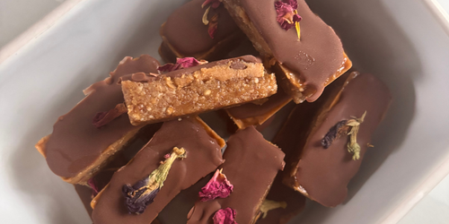 Caramel seed cycling + collagen chocolate slices with a dark chocolate top layer, a creamy cashew caramel middle, and an almond butter seed-based base, cut into small squares and topped with edible dried flowers.