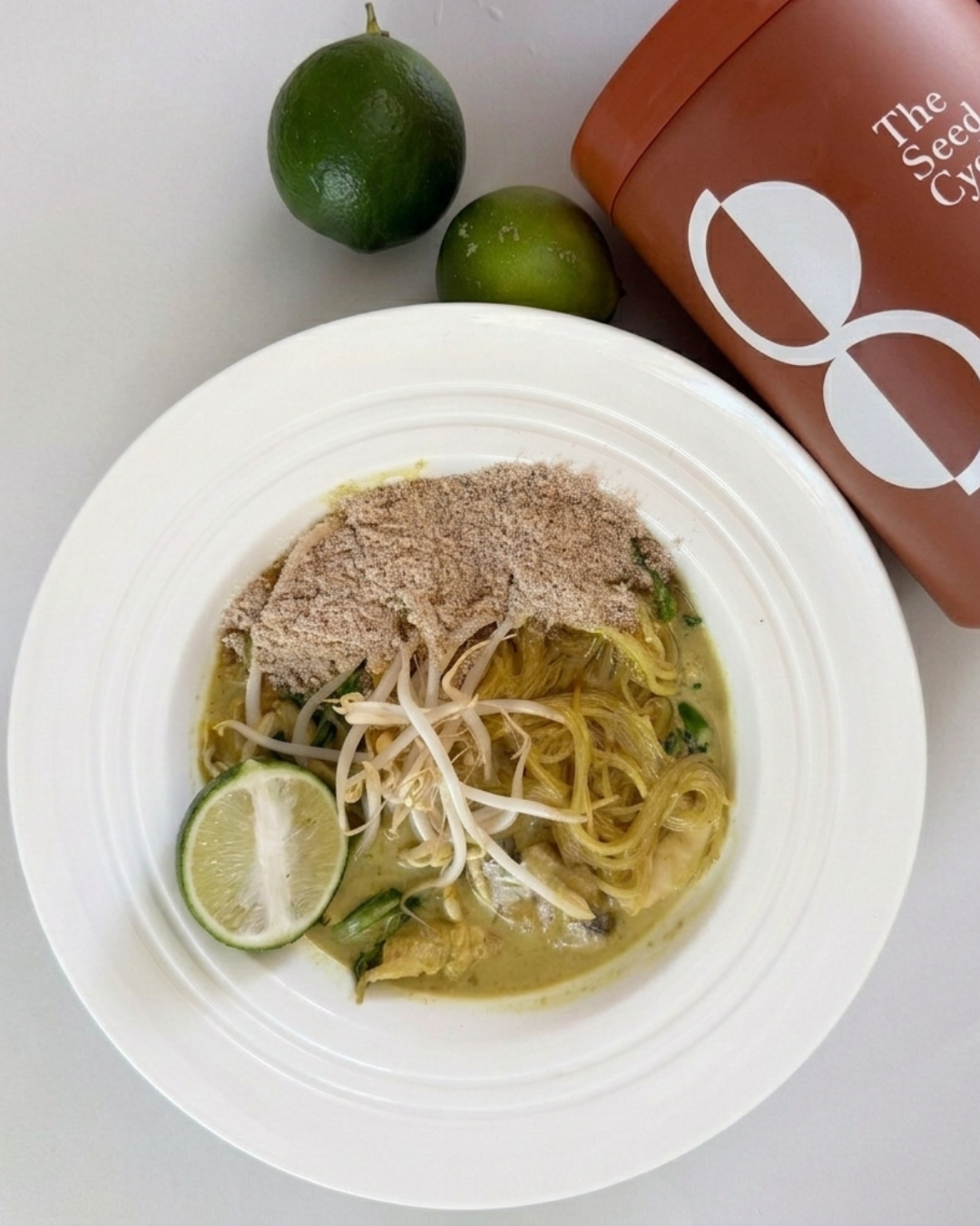 Gourmet green curry noodle bowl with "The Seed Cycle" terracotta canister and fresh limes