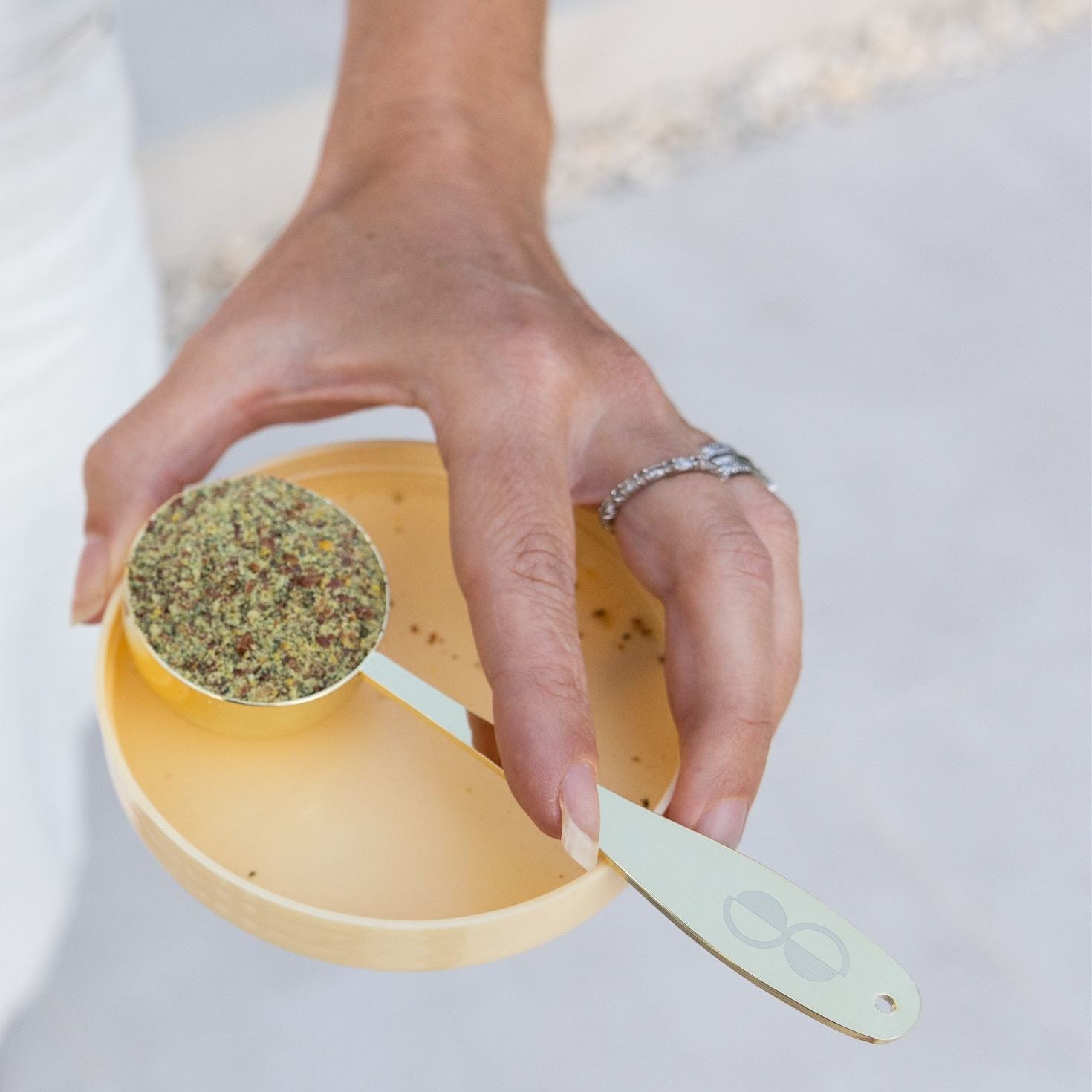 Hand holding a yellow measuring cup filled with herbs on a light background