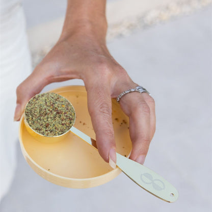 Hand holding a yellow measuring cup filled with herbs on a light background